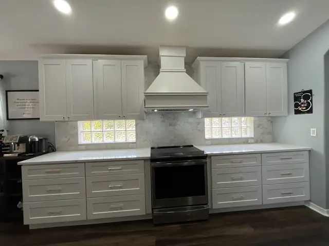 Kitchen range wall with white shaker cabinets, a custom white hood, marble-look backsplash and glass block windows